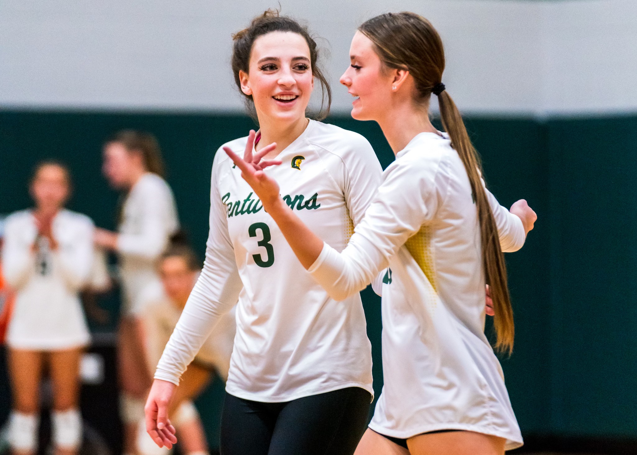 Two MCC Volleyball players talk to eachother on the court during a match with smiles on their faces.