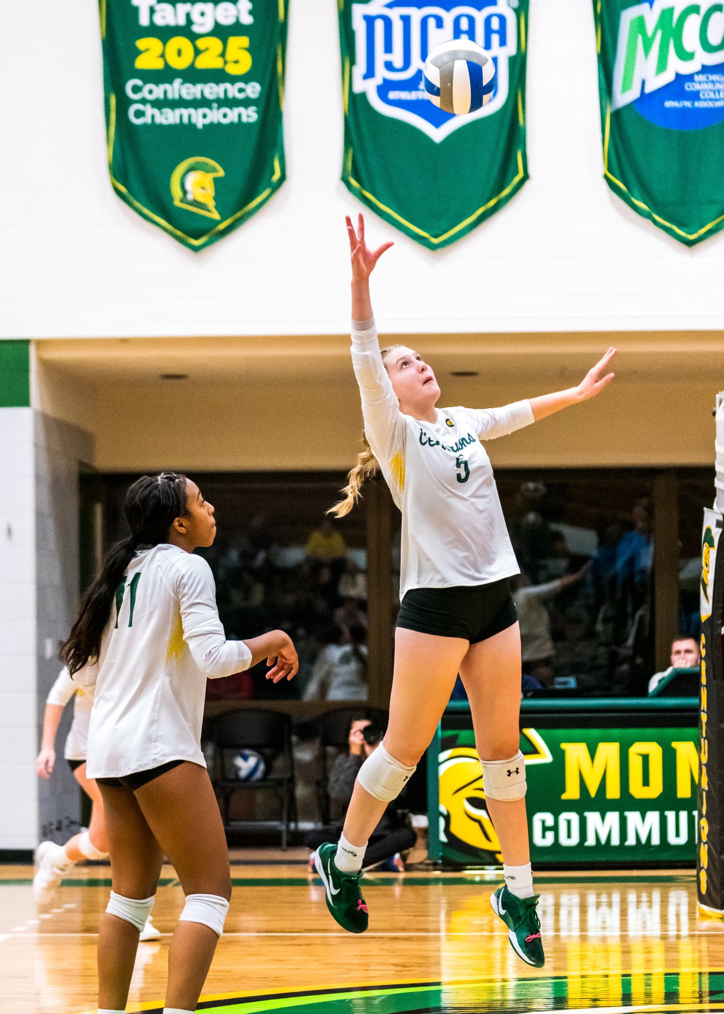 MCC volleyball player jumping to hit the ball on the court and another volleyball at the ready next to her to assist.